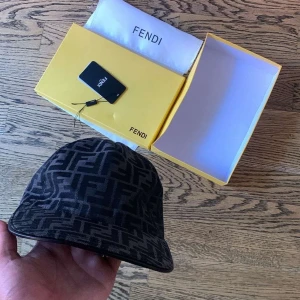 Svart Fendi keps med FF-mönster - Exklusiv svart keps från Fendi med ikoniskt FF-monogram i mörkgrått över hela kepsen. Justerbar rem baktill i skinn med Fendi-logga. Snygg böjd skärm och klassisk form, perfekt för dig som vill sticka ut med lyxig streetstyle.