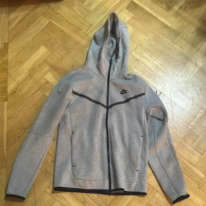 Grå Nike hoodiejacka med dragkedja - Snygg grå hoodiejacka från Nike med hel dragkedja och svart logga på bröstet. Jackan har huva, svarta detaljer och praktiska fickor framtill. Perfekt för chill eller träning, och den är tillverkad i mjukt syntetmaterial för skön känsla.