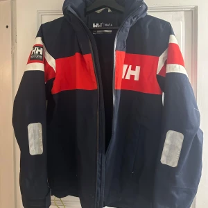 Helly Hansen marinblå vindjacka - Snygg marinblå vindjacka från Helly Hansen med röda och vita detaljer samt reflexer på ärmarna. Jackan har huva med gul insida. Den är i strl 164 vilket jag skulle säga är ca xs-s. Köpt förra året för 1660 och säljer för 350.