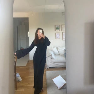 Svart långklänning med slits och öppen rygg  - Svart långklänning med vida ärmar och snygg slits Klänningen har en djup v-ringning i ryggen som ger en cool touch. Perfekt för dig som gillar minimalistisk stil med en twist. Materialet är mjukt och följsamt.