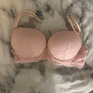 Två spetsiga bh:ar i rosa och rött i strl XS - Säljer två snygga bh-ar i storlek XS. En är ljusrosa med blommig spets, vadderade kupor och justerbara axelband. Den andra är röd med blommig spets, rosett framtill och justerbara axelband. Båda har fina spetsdetaljer och ger en feminin look.