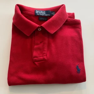 Röd pikétröja från Polo Ralph Lauren - Klassisk röd pikétröja från Polo Ralph Lauren med blå broderad logga på bröstet. Tröjan har krage, två knappar och korta ärmar. Tillverkad i bomull för en skön och luftig känsla. Perfekt för en clean och sportig stil.