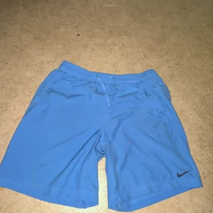 Blå Nike shorts  - Snygga blå shorts från Nike med elastisk midja och snörning framtill. Klassisk design med två sidofickor och diskret Nike-logga på benet. Perfekta för träning eller chill dagar. Lätta och bekväma i syntetmaterial.