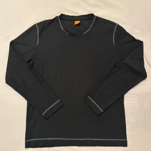 Svart långärmad tröja från Hugo Boss - Svart långärmad tröja från Hugo Boss med kontrastsömmar i grått. Tillverkad i 100% bomull för en skön och avslappnad känsla. Klassisk rund halsringning och diskret logga på ryggen. Perfekt för en stilren och enkel look.