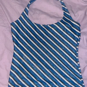 Blå glittrig randig halterneck topp - Säljer en snygg halterneck topp med blå, svarta och vita diagonala ränder och glittrigt tyg. Perfekt för dig som vill sticka ut med lite extra shimmer. Toppen är ärmlös och har en tight passform som framhäver axlarna.