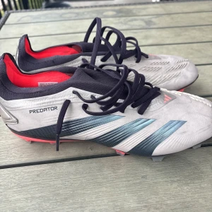 Adidas Predator fotbollsskor grå/svart - Snygga Adidas Predator fotbollsskor i grått med svarta och blå detaljer samt röda inslag. Skorna har snörning, mönstrad ovandel och en yttersula med dobbar för bra grepp på planen. Insidan är fodrad i rött och designen är modern och sportig.