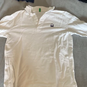 Vit pikétröja från Benetton - Klassisk vit pikétröja från Benetton med broderad logga på bröstet. Tröjan har krage, knappar framtill och är tillverkad i mjuk bomull. Perfekt för en clean och stilren look. Storlek: 155 cirka, skick: 9/10, kontakta vid frågor!