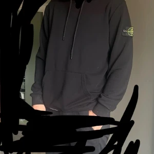 Svart hoodie från Stone Island - Svart hoodie från Stone Island med klassisk patch på vänster ärm. Tröjan har huva med snörning och en stor magficka framtill. Mjuk bomullskänsla och relaxed fit, perfekt för dig som gillar streetwear.