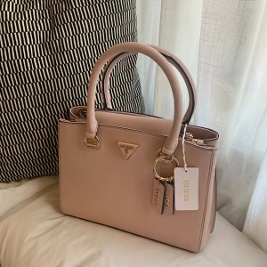 Ljusrosa handväska från Guess - Säljer en assnygg oanvänd ljusrosa märkesväska från Guess med gulddetaljer. Väskan har två rejäla handtag, och dragkedja baktill och kommer med dustbag och matchande axelrem.