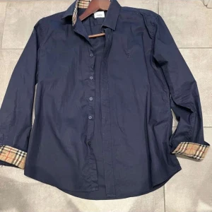 Mörkblå skjorta från Burberry - Snygg mörkblå skjorta från Burberry med klassiskt rutigt tyg på insidan av kragen och ärmsluten. Skjortan har knappar framtill och lång ärm, perfekt för en stilren look. Diskret broderad logga på bröstet.