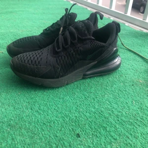 Svarta Nike Air Max 270 sneakers - Säljer ett par helsvarta Nike Air Max 270 sneakers med mesh-ovandel och synlig Air-enhet i hälen. Skorna har snörning och en sportig vibe, perfekta för dig som gillar streetstyle. Snygg och bekväm modell med rund tå och platt sula.