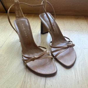 Beige sandaler med klack från Nine West - Snygga beige sandaler från Nine West med smala remmar och fyrkantig tå. Skorna har hög klack och justerbart spänne i guld. Tillverkade i skinn. (Observera liten slitning på banden) 