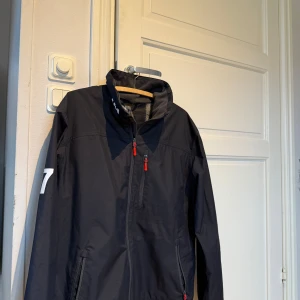 Svart vindjacka Helly Hansen L - Svart vindjacka från Helly Hansen med vita HH-detaljer på axeln och röd dragkedja. Jackan har hög krage, meshfoder och flera fickor med dragkedja. Perfekt för blåsiga dagar och snygg till jeans eller träningsbyxor.