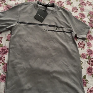 Armani tshirt  - Snygg grå t-shirt från Emporio Armani med svart och mörkgrå randdetalj över bröstet och liten logga. Klassisk rund hals och korta ärmar. Perfekt för en clean och stilren look. 