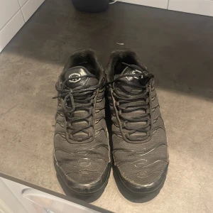 Svarta Nike Air Max Plus TN sneakers - Säljer ett par svarta Nike Air Max Plus TN sneakers med klassisk vågig design och TN-logga på hälen och sulan. Skorna har snörning, mesh- och syntetmaterial samt en chunky sula med synliga luftbubblor. Perfekta för dig som gillar streetwear och sportig stil.