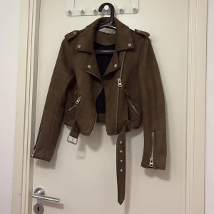 Brun mocka bikerjacka från Zara - Snygg brun bikerjacka i mocka från Zara med coola silvriga dragkedjor, axelklaffar och bälte i midjan. Jackan är croppad och har långa ärmar med dragkedjor vid ärmsluten. Perfekt för dig som vill ha en edgy look. Pris kan diskuteras 