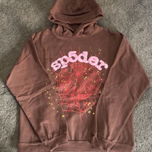 Sp5der hoodie brun - Säljer en brun hoodie med en cool spindelnätsdesign i rött och texten 'sp5der' i rosa på framsidan. Perfekt för en avslappnad stil med en unik touch. Hoodien har långa ärmar och en klassisk huva.