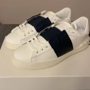 Vit och marinblå sneaker - Snygga vita sneakers med en bred marinblå rem över snörningen. Skorna har en stilren design med rund tå och är perfekta för en avslappnad look.
