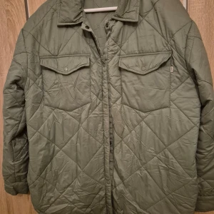 Grön quiltad jacka från CRW - Säljer en grön quiltad jacka från CRW. Jackan har en klassisk krage och knäppning framtill. Två bröstfickor med lock och knappar ger en stilren look. Perfekt för kyligare dagar.