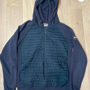 Colmar cardigan - Säljer en snygg mörkblå hoodie från Colmar  med quiltad front och dragkedja. Den har en huva med snörning och långa ärmar. Perfekt för en casual look. Perfekt till våren/sommarkvällar.