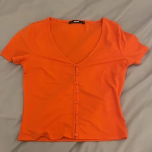 Orange kortärmad topp från BikBok - Säljer en snygg orange topp från BikBok med korta ärmar och v-ringning. Toppen har knappar framtill som ger en stilren look. Mjukt material 💘