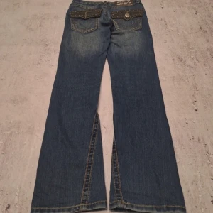 Blå jeans med vida ben från True Religion - Snygga blå jeans från True Religion med vida ben och coola detaljer som nitar på fickorna. De har en klassisk femficksdesign och en knappgylf. Perfekta för en avslappnad stil med en touch av edge.saknar en knapp men inget man tänker på