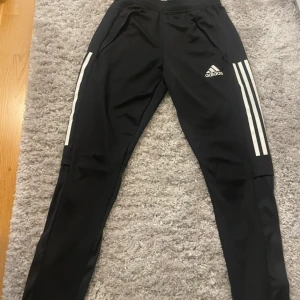 Svarta träningsbyxor från Adidas - Snygga svarta träningsbyxor från Adidas med de klassiska tre vita ränderna längs sidorna. Byxorna har en smal passform och är perfekta för sport och träning. De har även praktiska fickor och en elastisk midja för extra komfort.