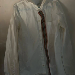 Vit skjorta från Burberry - Säljer en stilren vit skjorta från Burberry med en unik randig detalj i rött, svart och beige längs knappraden. Skjortan har långa ärmar och klassiska knappar framtill. Perfekt för en elegant look.