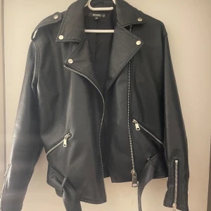 Svart skinnjacka från Bershka - Snygg svart skinnjacka från Bershka med coola dragkedjedetaljer och bälte i midjan. Perfekt för en edgy look. Jackan har långa ärmar och en klassisk bikerstil med knappar på kragen. Köpare står för frakt