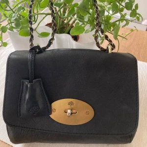 Svart Mulberry lilly axelväska med kedja, äkta skinn - Snygg svart axelväska i skinn med guldfärgad kedja och låsdetalj. Väskan har en elegant design med en praktisk insida och dragkedja. Perfekt för att ge din outfit en stilfull touch.