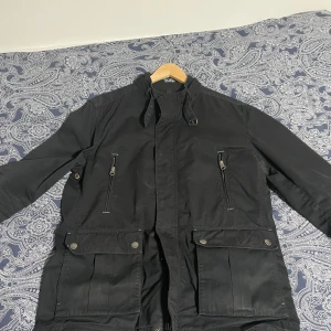 Svart field jacket från Massimo dutti  - Snygg svart parkajacka från Massimo dutti med dragkedja och knappar. Jackan har flera praktiska fickor med dragkedjor och knappar, perfekt för att hålla dina saker säkra. Den har en stilren design och är långärmad, vilket gör den idealisk för kyligare väder.