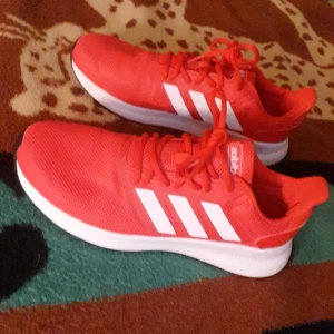 Röda sneakers från Adidas - Snygga röda sneakers från Adidas med vita detaljer och snörning. Perfekta för en sportig look med de klassiska tre ränderna på sidan. Skorna har en bekväm passform och en vit sula som ger bra stöd.