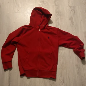 Röd ralph lauren hoodie med dragkedja - Säljer en snygg ralph lauren röd hoodie med dragkedja och huva. Den har en liten broderad logga på bröstet och långa ärmar. Perfekt för en avslappnad stil.