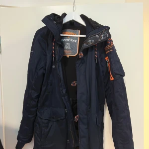 Mörkblå parkajacka från Superdry - Säljer en snygg mörkblå parkajacka från Superdry med orange detaljer. Jackan har en huva med snörning och flera fickor, inklusive en med dragkedja på ärmen. Perfekt för kyliga dagar med sin MicroFibre-isolering.