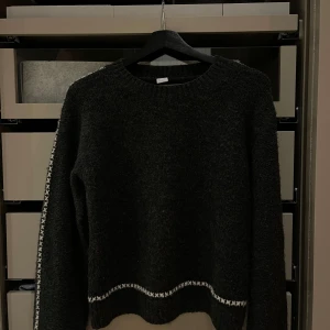 Mörkgrå stickad tröja med mönster - Säljer en mysig mörkgrå stickad tröja med vita mönster längs ärmarna och nederkanten. Perfekt för kyliga dagar och ger en stilren look. Tröjan har långa ärmar och en rund halsringning.