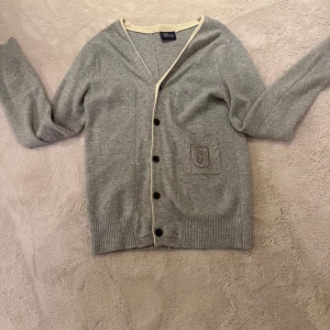 Grå kofta från Peak Performance - Säljer en stilren grå kofta från Peak Performance med knappar framtill och en broderad detalj på fickan. Koftan är använd men i väldigt bra skick inga skador eller fläckar den ser ut som ny. Den är i st M 