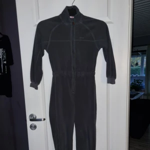 Grå fleeceoverall med dragkedja - Mysig grå fleeceoverall med dragkedja framtill. Perfekt för kalla dagar med sin varma och bekväma design. Overallen har långa ärmar och ben med muddar för extra komfort.