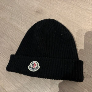 Svart  mössa från Moncler - Snygg svart ribbad mössa från Moncler med broderad logotyp framtill. Perfekt för kyliga dagar och ger en stilren look. 