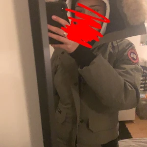 Grön parkajacka med pälsdetalj - Säljer en grön parkajacka från Canada Goose med pälsdetalj på huvan. Jackan har en klassisk design med dragkedja och knappar framtill med två uppe och två fickor nere . Perfekt för kalla dagar.