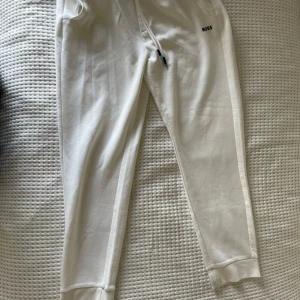 Vita mjukisbyxor från Boss - Snygga vita mjukisbyxor från Boss med elastisk midja och snörning. Byxorna har en avslappnad passform och ribbade muddar vid bensluten. Diskret Boss-logga framtill och en större logga på bakfickan.