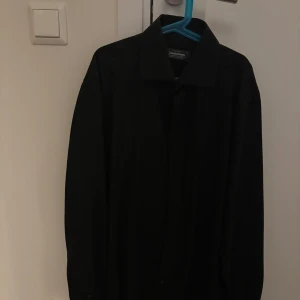 Svart skjorta från Dressmann - Snygg svart skjorta från Dressmann, perfekt för en stilren look. Skjortan har en klassisk krage och är långärmad. Materialet är skrynkelfritt vilket gör den lättskött och alltid redo att användas.
