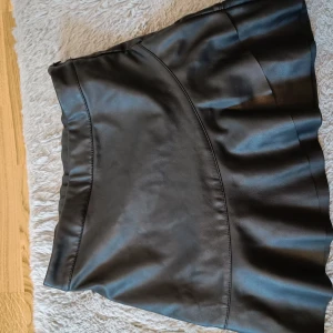 Svart skinnkjol från KappAhl - Snygg svart skinnkjol från KappAhl i storlek 152. Kjolen har en dragkedja på sidan för enkel på- och avtagning. Perfekt för en stilren look.