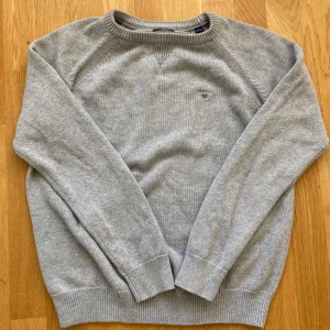 Grå stickad tröja från Gant - Säljer en stilren grå stickad tröja från Gant med ett diskret broderat emblem på bröstet. Tröjan har långa ärmar och ribbade muddar vid ärmslut och nederkant. Perfekt för kyligare dagar.