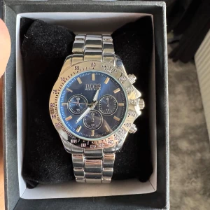 Silverfärgad klocka från Boss - Snygg och stilren klocka från Boss med en silverfärgad länkarmband och blå urtavla. Klockan har tre mindre urtavlor och en kronografdesign. Perfekt för den som vill ha en elegant accessoar.