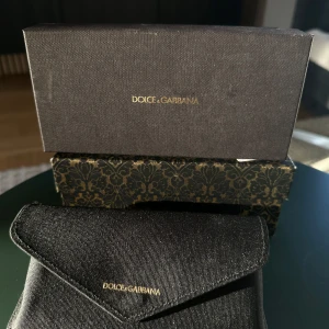 Svart glasögonfodral från Dolce & Gabbana - Elegant svart glasögonfodral från Dolce & Gabbana med guldlogotyp. Fodralet har en stilren design med ett praktiskt magnetlås. Perfekt för att skydda dina glasögon med stil. Säljer då de ändats står och tar plats.  Kartongerna är ju fina själva bara som det är men sedan har du det praktiska solglasögon fodralet. Vill tillägga att det aldrig är använt är dock lite dammigt på bilden. Pris går att diskuteras.