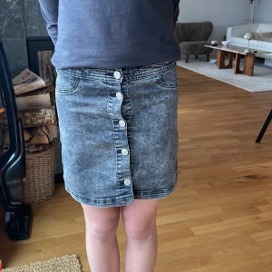 Grå jeanskjol från H&M - Snygg grå jeanskjol från H&M med knappar framtill. Kjolen har en klassisk design med en medel längd. Den är i bra skick! Skulle säga att den är ganska stor i storleken? Jag är 13 och får på mig den. 