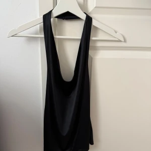 Svart ärmlös topp - Snygg svart ärmlös topp i ett glansigt material. Perfekt för en stilren look. Toppen har en enkel design med en djup urringning och breda axelband.