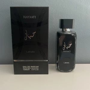 Hayaati Eau de Parfum från Lattafa - Elegant och mystisk parfymflaska från Lattafa, Hayaati. Flaskan är svart med stilrena linjer och har en matchande svart kork. Förpackningen är också svart med dekorativa mönster och text i silver. Innehåller 100 ml av en doft som utstrålar lyx och sofistikation.