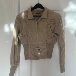 Beige jacka från Liz Fashion - Snygg beige jacka från Liz Fashion med dragkedja och knappar. Jackan har en croppad design med ribbade detaljer vid midjan och ärmsluten. Perfekt för en stilren look!