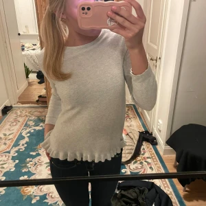 Grå tröja med volangdetalj - Säljer en stilren grå tröja med volangdetalj vid nederkanten. Tröjan har trekvartsärmar och en rund halsringning. Perfekt för en avslappnad look.